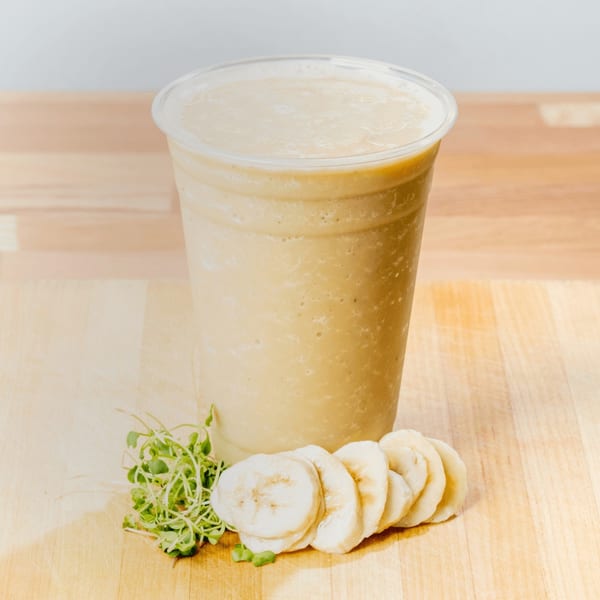 A banana smoothie in a clear plastic cup, with sliced bananas and microgreens on a wooden surface.