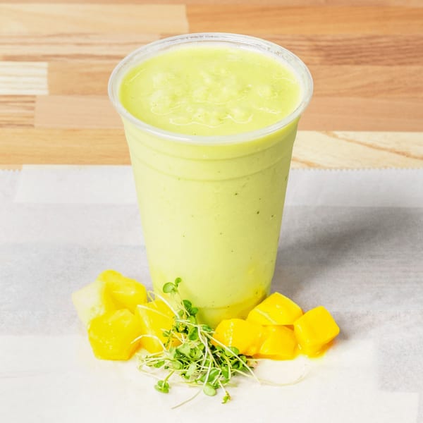 A vibrant green smoothie in a clear cup, surrounded by fresh mango chunks and microgreens.