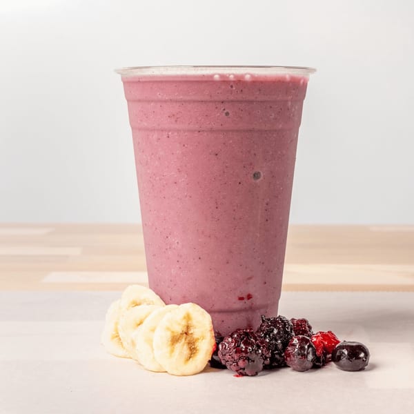 Pink berry smoothie in a clear cup with banana slices and mixed berries on a white surface.