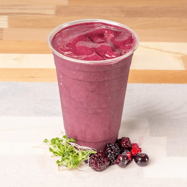 Berry smoothie in a clear cup with fresh berries and microgreens on a white surface.