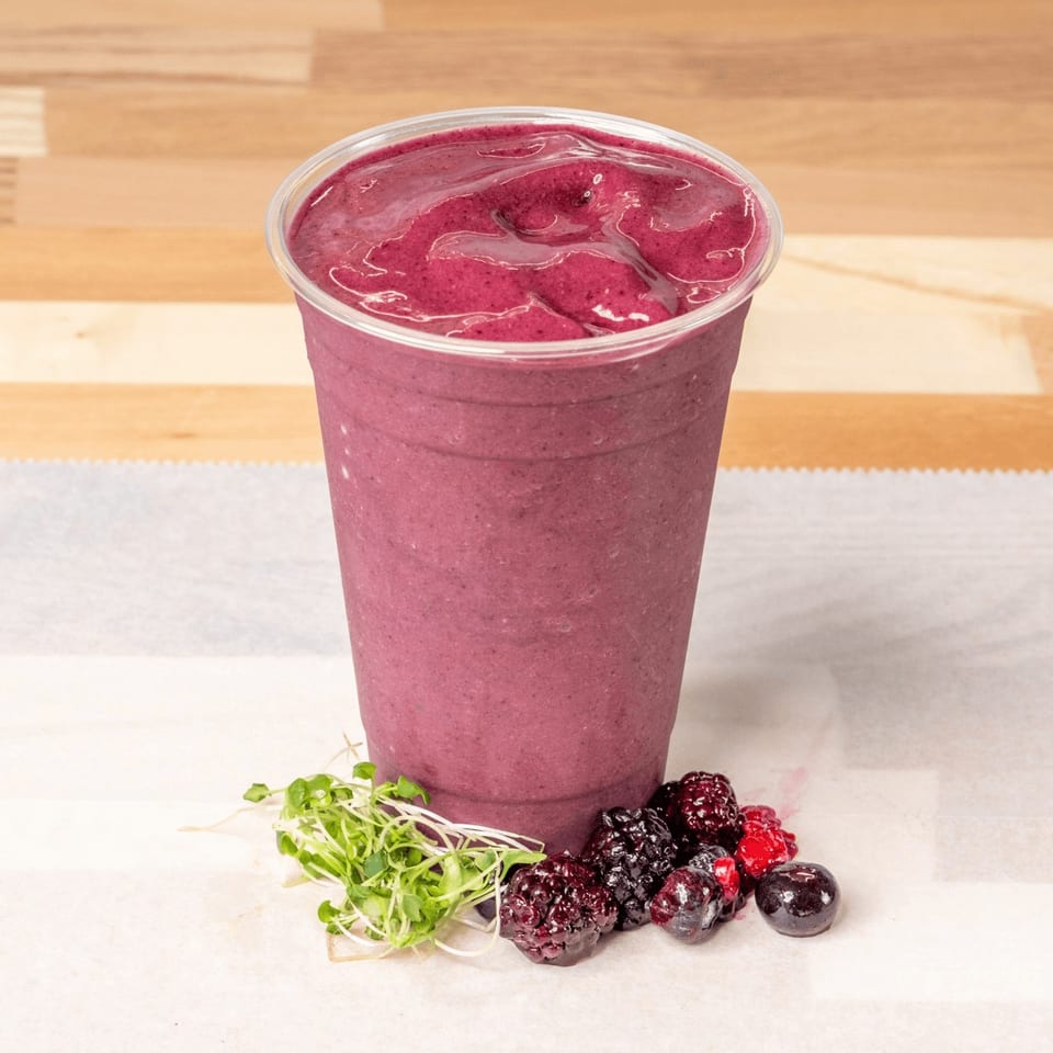 Berry smoothie in a clear cup with fresh berries and microgreens on a white surface.