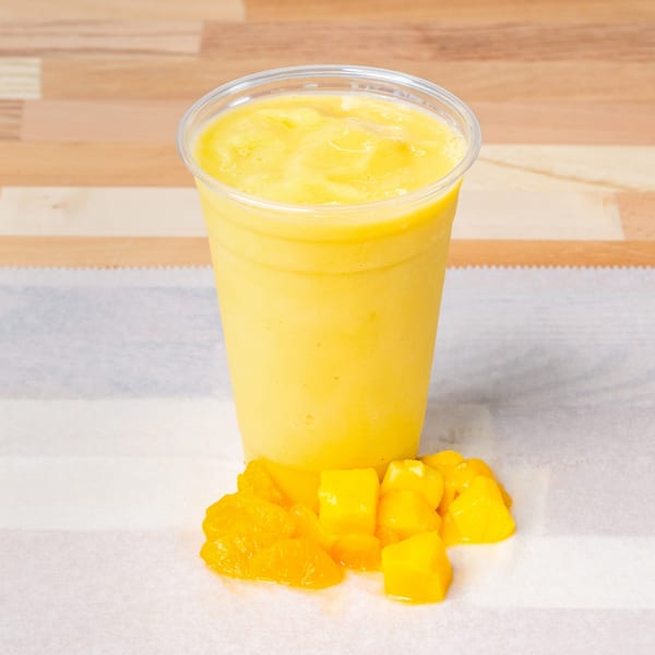 A vibrant yellow mango smoothie in a clear plastic cup, with fresh mango chunks beside it.